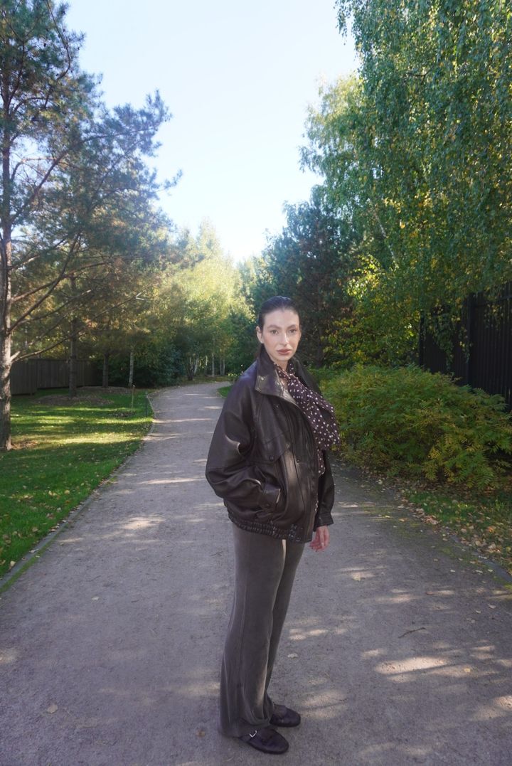 An oversized brown leather jacket over a polka-dot scarf and velvet trousers on a garden path